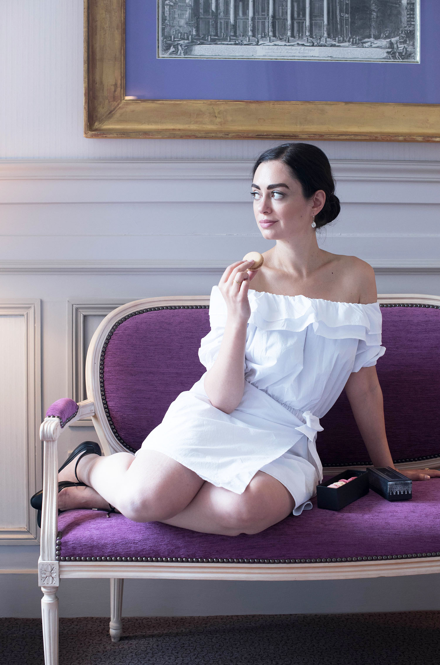 Cee Fardoe, the fashion blogger behind Coco & Vera, sits on a Louis XVI bench eating Laduree macarons while wearing a white dress from REVOLVE