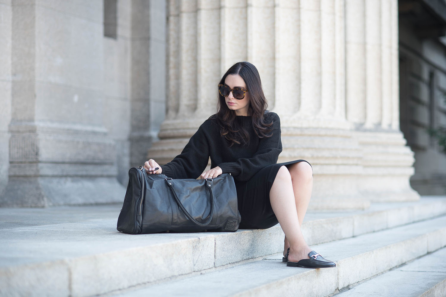 Winnipeg fashion blogger Cee Fardoe sits on a set of stairs wearing a black Forever 21 dress and holding a Mahi Leather duffle bag