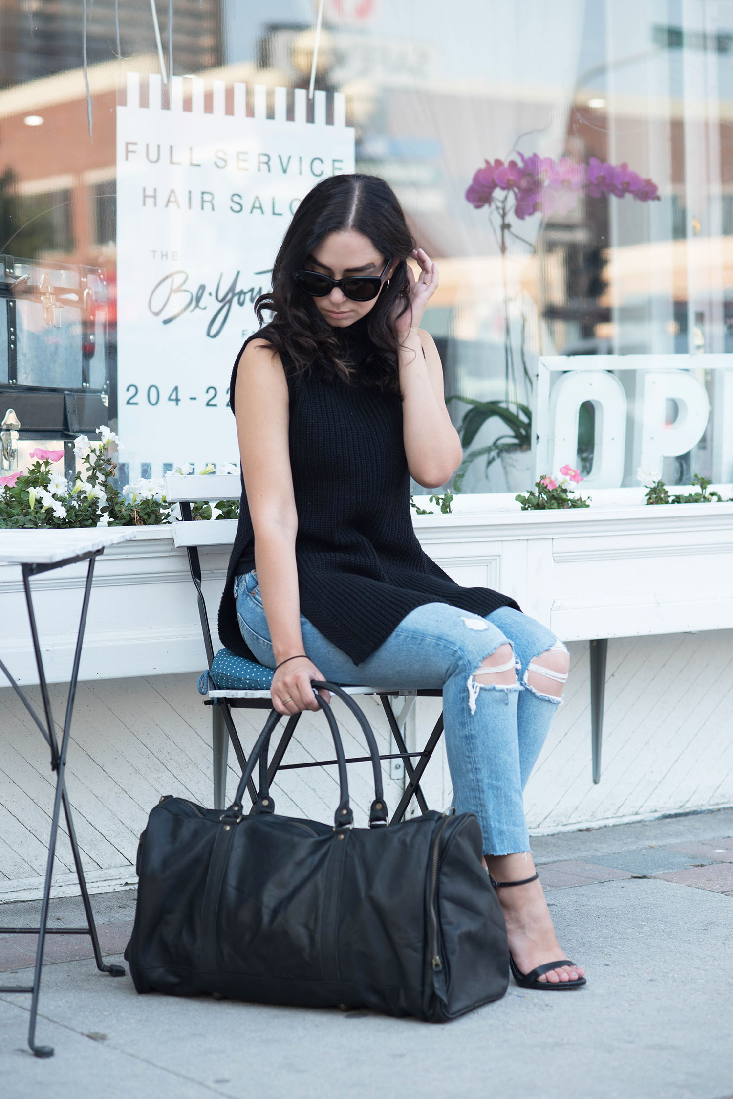 Winnipeg fashion blogger sits in front of a white cafe carrying a Mahi Leather duffle bag and wearing an Aritzia sweater