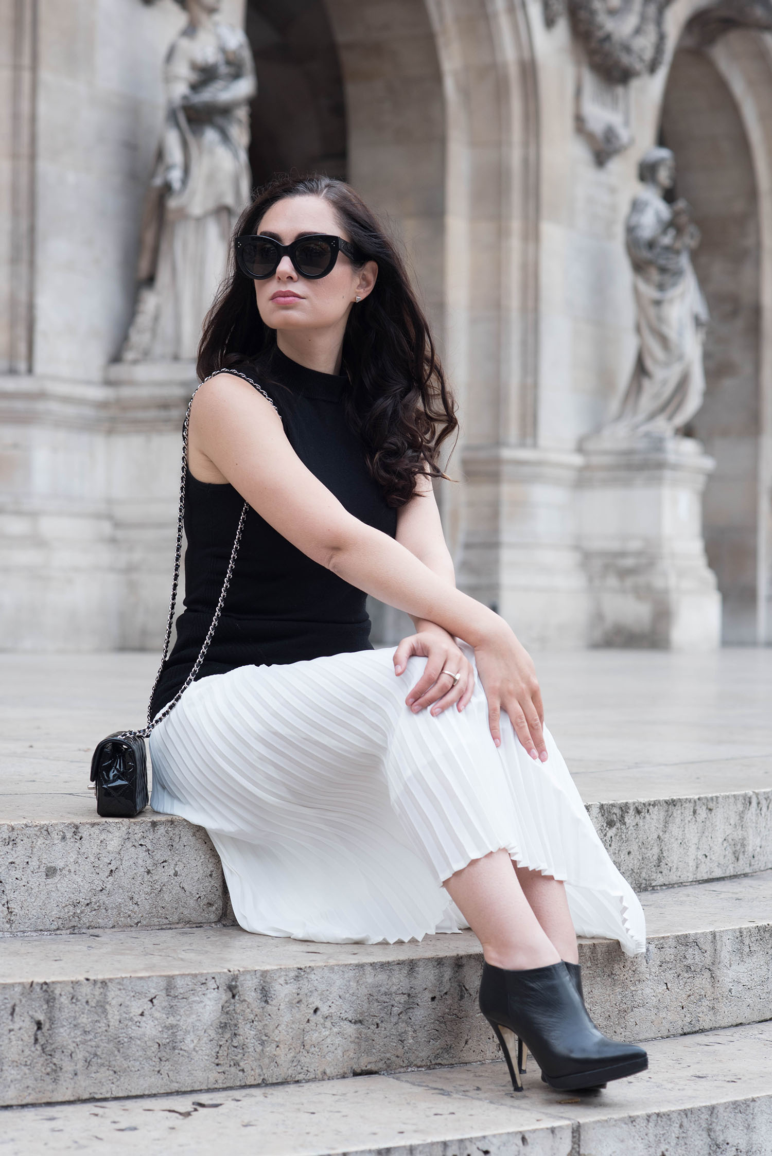 Winnipeg fashion blogger Cee Fardoe of Coco & Vera sits on the stairs of the Opera Garner in Paris, wearing an Aritzia pleated skirt, Celine Audrey sunglasses and a Chanel 2.55 handbag