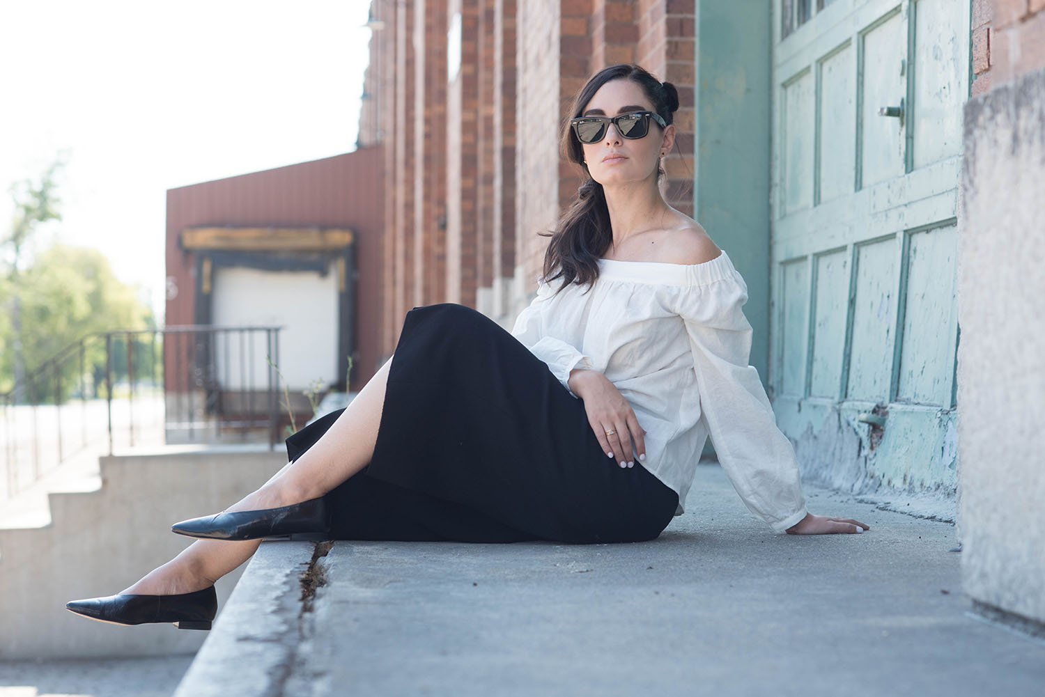 Fashion blogger Cee Fardoe of Coco & Vera sits in an old loading bay wearing a L'Academie off-shoulder blouse, RayBan Wayfarer sunglasses and black Aritzia culottes