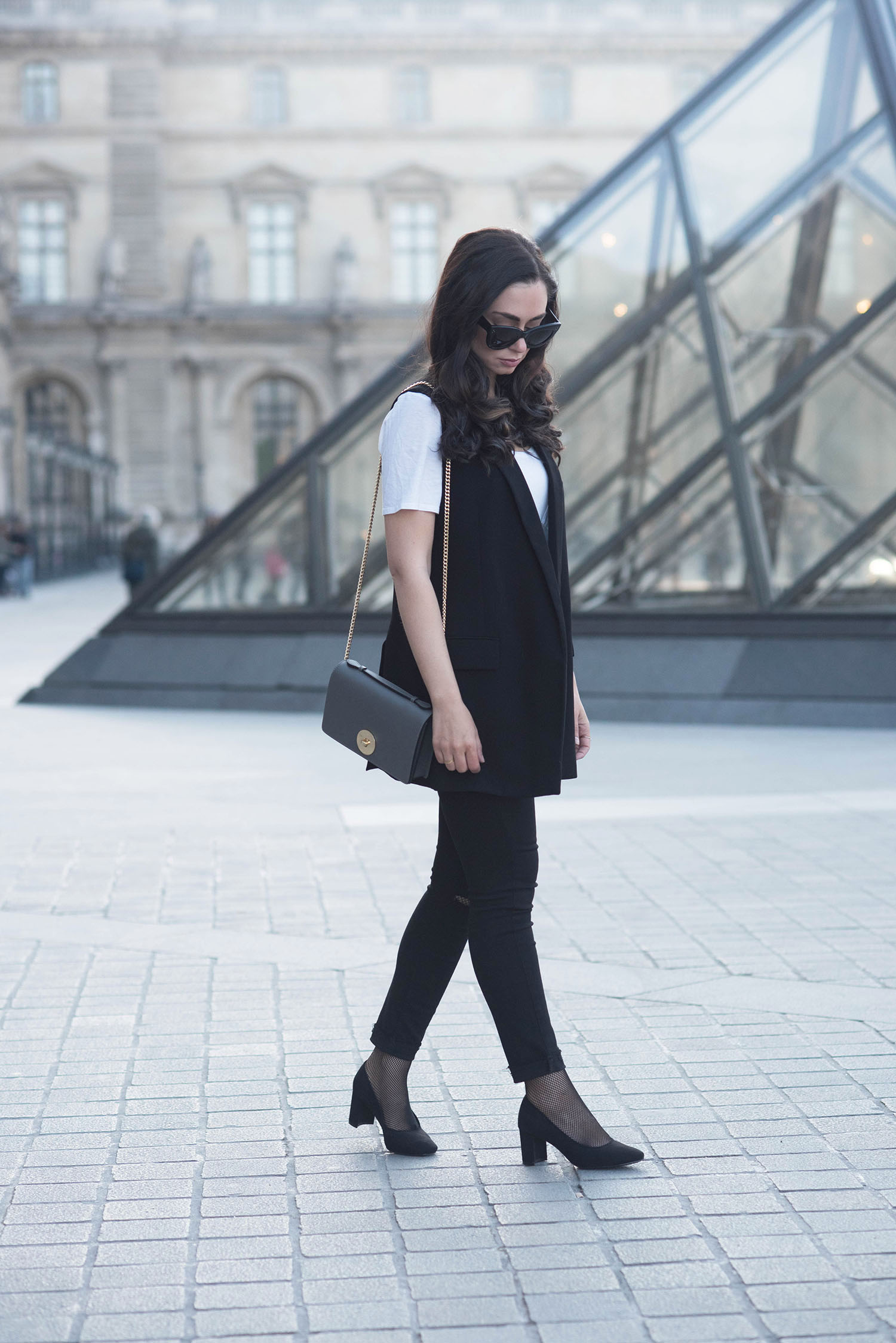 Fashion blogger Cee Fardoe of Coco & Vera walks at the Louvre museum wearing a black Zara waistcoat and carrying a grey Camelia Roma crossbody bag