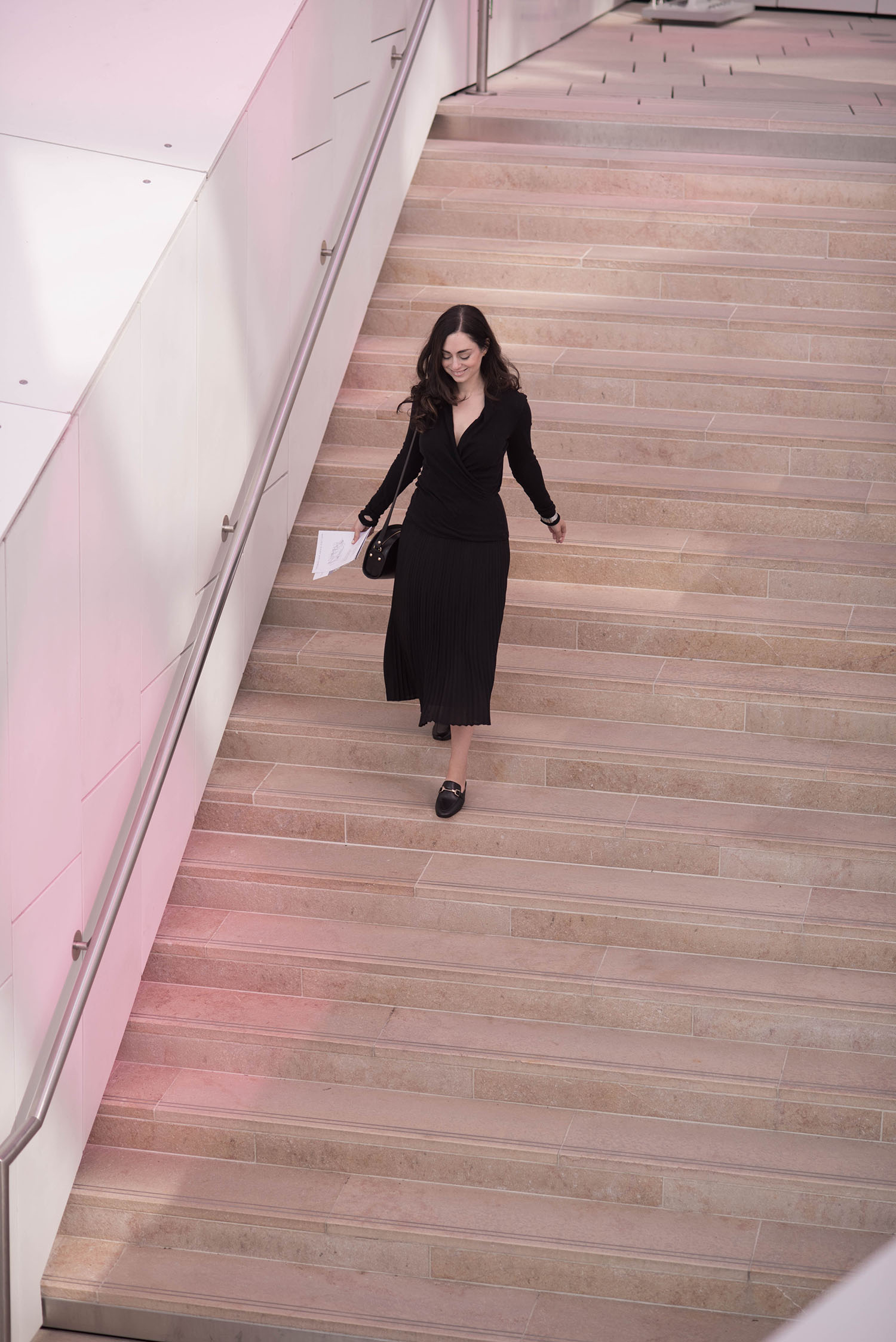 Fashion blogger Cee Fardoe of Coco & Vera walks down the stairs at the Fondation Louis Vuitton in Paris wearing an Aritzia pleated skirt and an Enza Costa wrap top