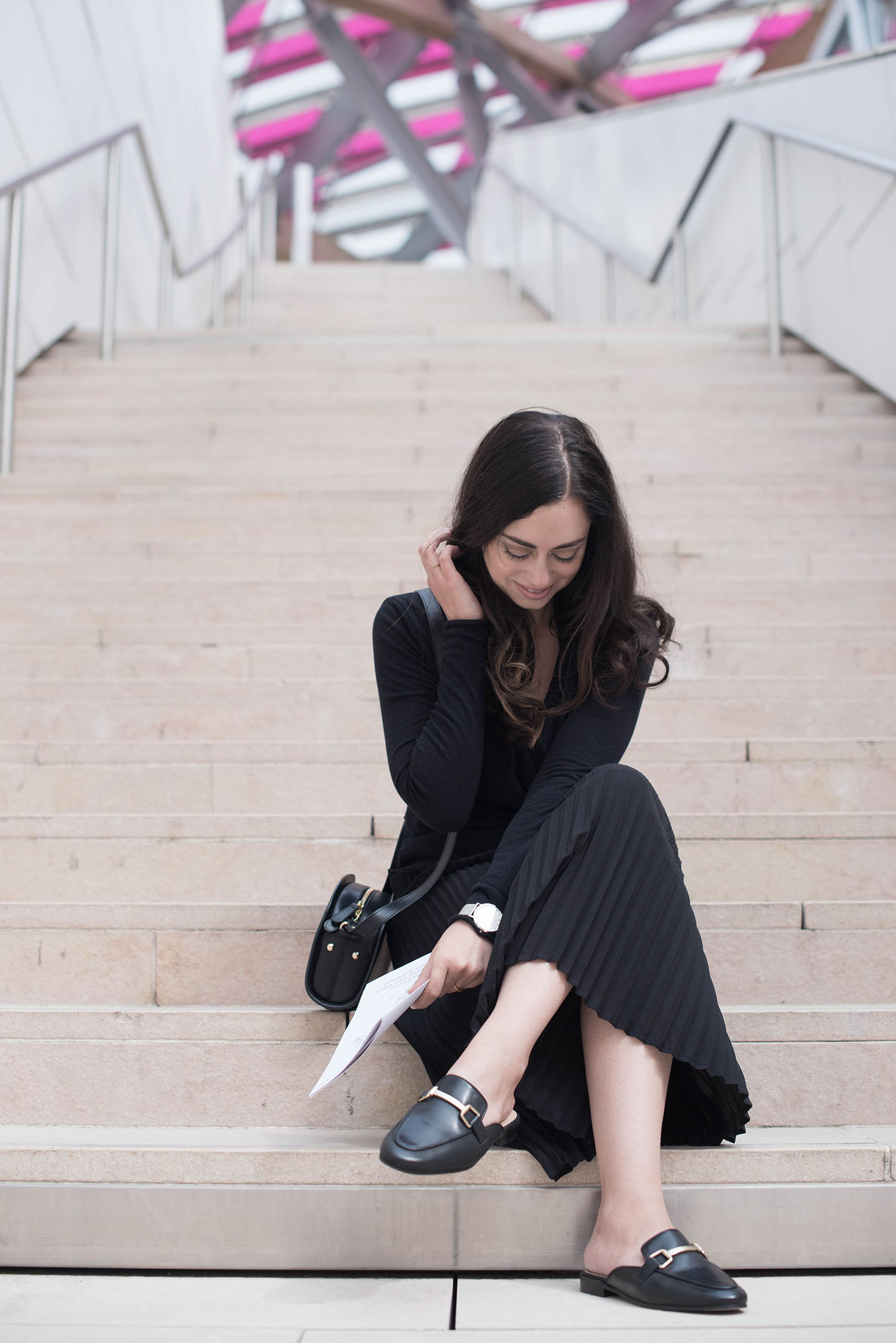 Fashion blogger Cee Fardoe of Coco & Vera sits on the stairs at the Fondation Louis Vuitton wearing an Aritzia pleated skirt and Jonak mules