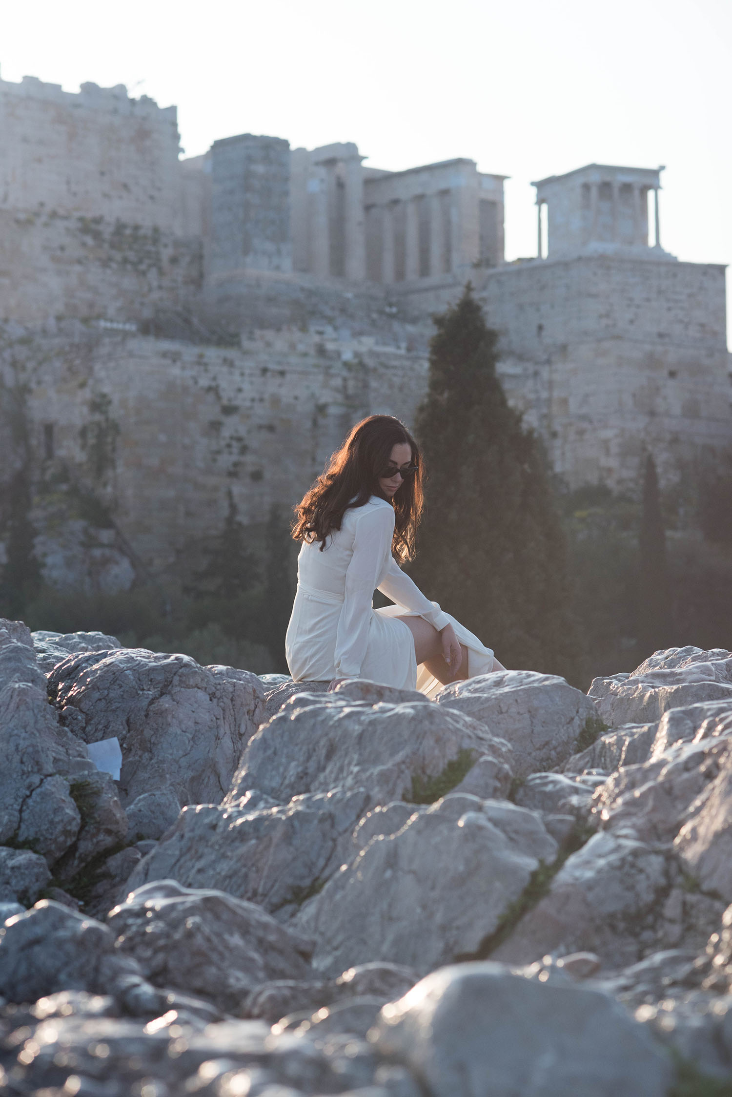 Canadian fashion blogger Cee Fardoe sits in front of Acropolis Hill in Athens, wearing a white maxi dress from Lovers + Friends and RayBan Wayfarer sunglasses
