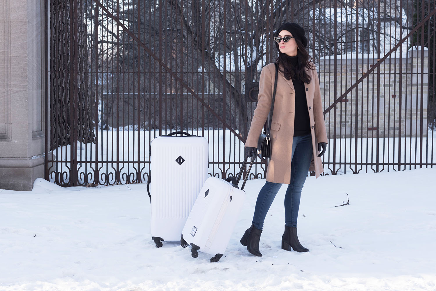 Winnipeg fashion blogger Cee Fardoe of Coco & Vera pulls her white Herschel Supply Co. suitcase through the snow wearing Aldo leather boots and an Anthropologie Bonnie beret
