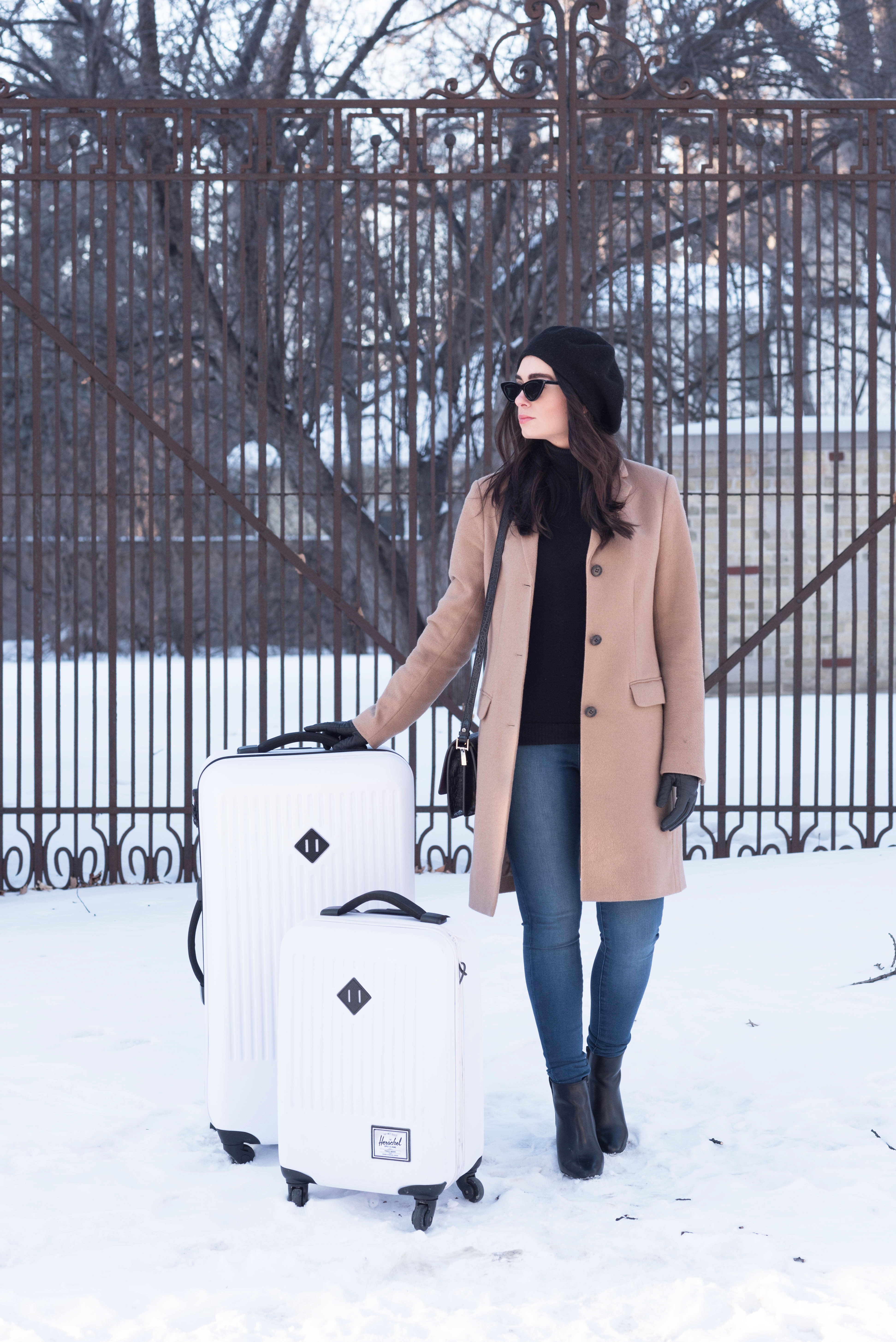 Fashion blogger Cee Fardoe of Coco & Vera stands in the snow next to her white Herschel Supply Co suitcases wearing a Uniqlo camel coat and Mott & Bow jeans