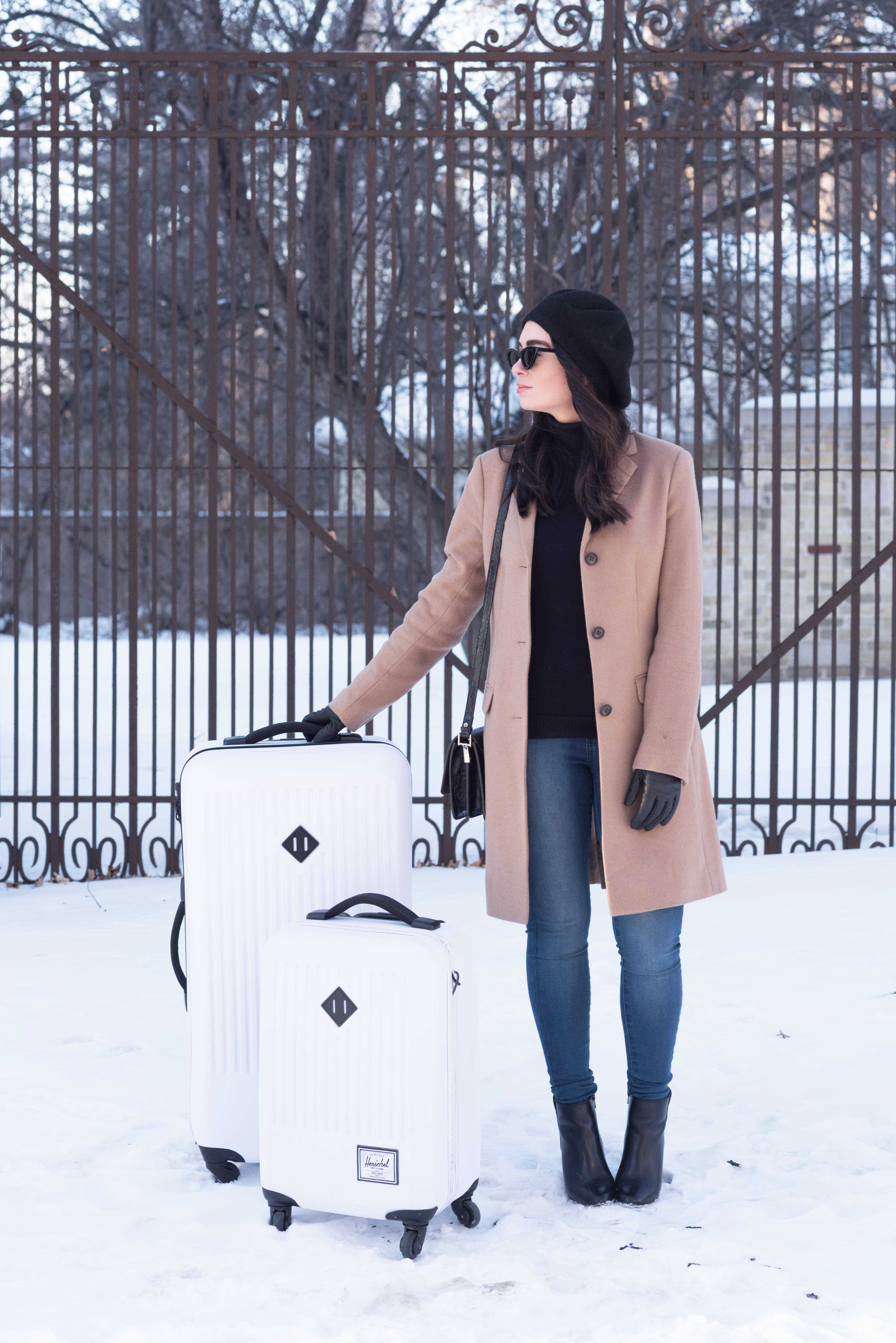 Fashion blogger Cee Fardoe of Coco & Vera leans on her white Herschel Supply Co. suitcase, wearing a Uniqlo camel coat and Le Chateau black sweater