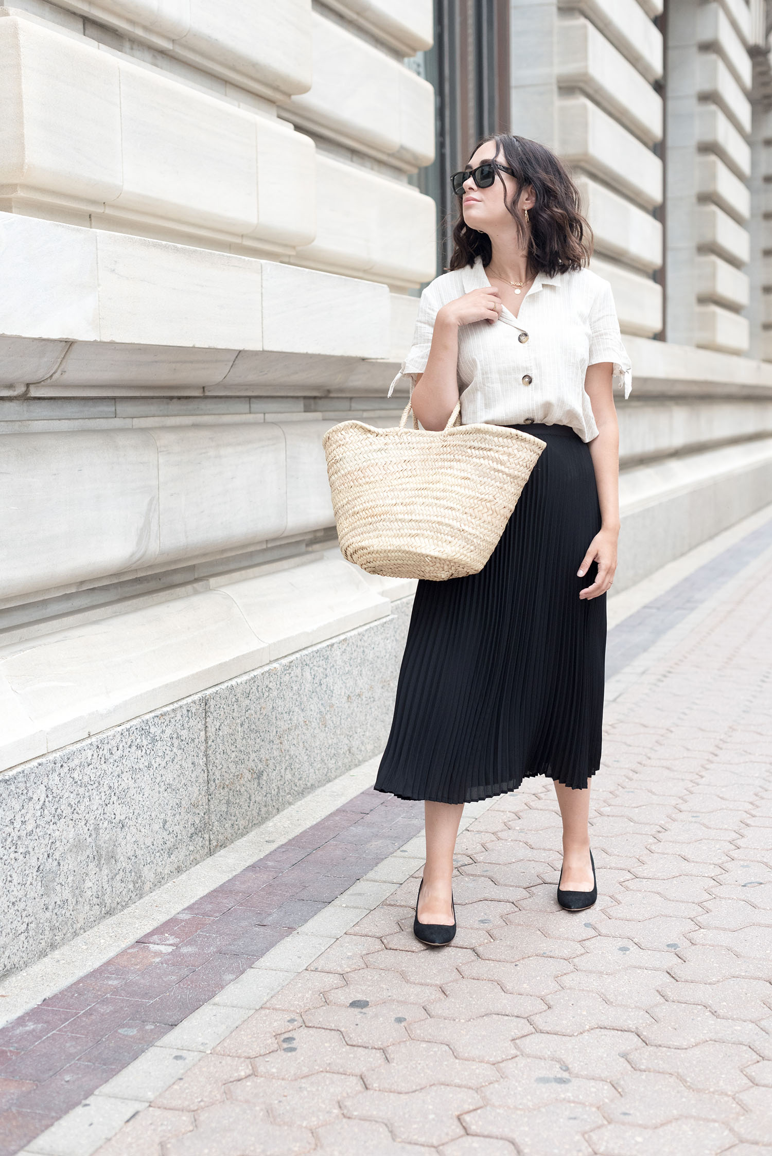 Top Winnipeg fashion blogger Cee Fardoe of Coco & Vera stands in the Exchange District wearing an Aritzia pleated skirt and carrying a Sezane straw tote bag