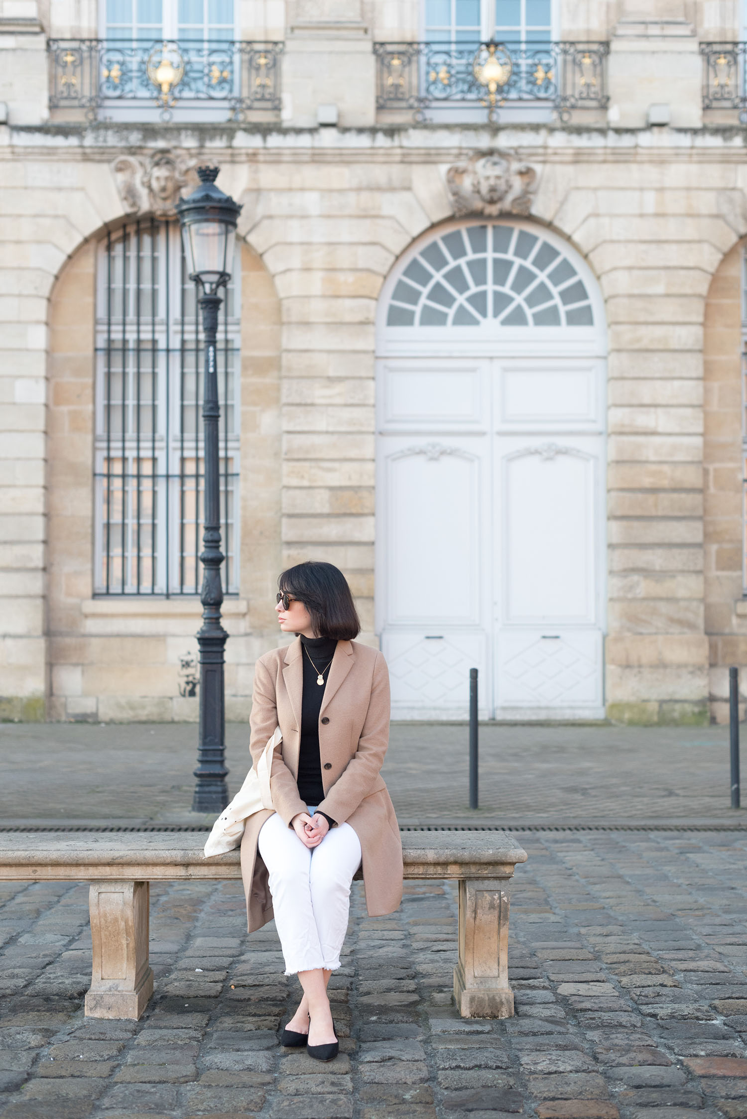 Top Winnipeg fashion blogger Cee Fardoe of Coco & Vera sits at Grande Place in Bordeaux, wearing a Uniqlo camel coat and Mavi white jeans