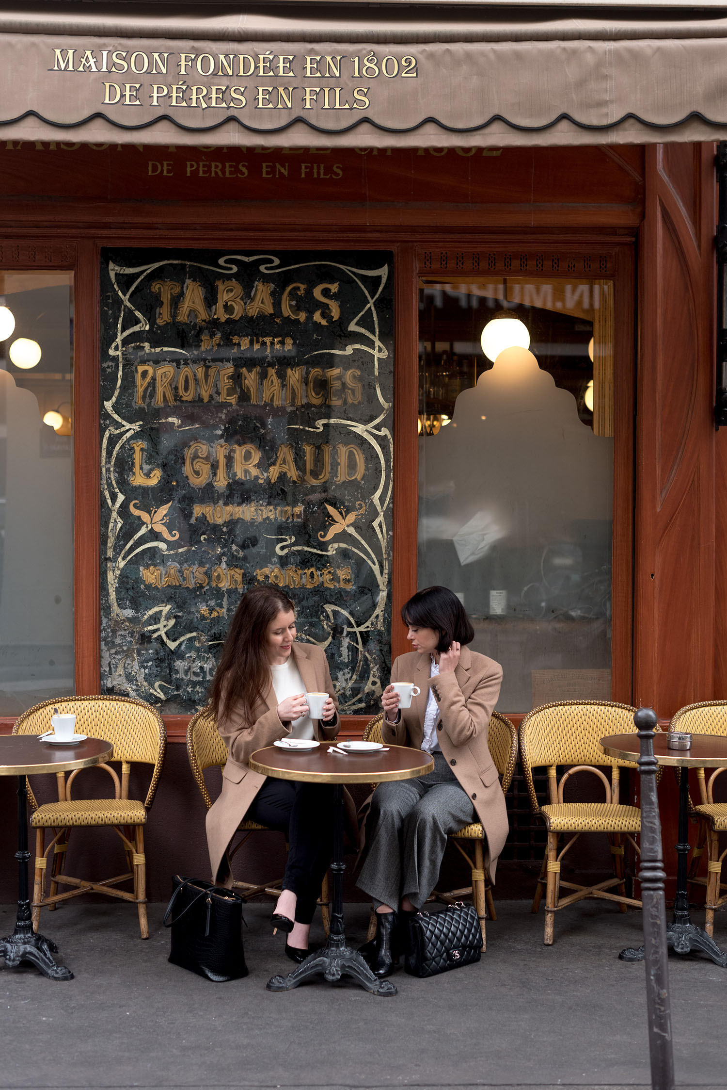 Top Canadian fashion bloggers Cee Fardoe of Coco & Vera and Lyndi Barrett of Style Calling sit at a Parisian cafe, wearing Uniqlo camel coats