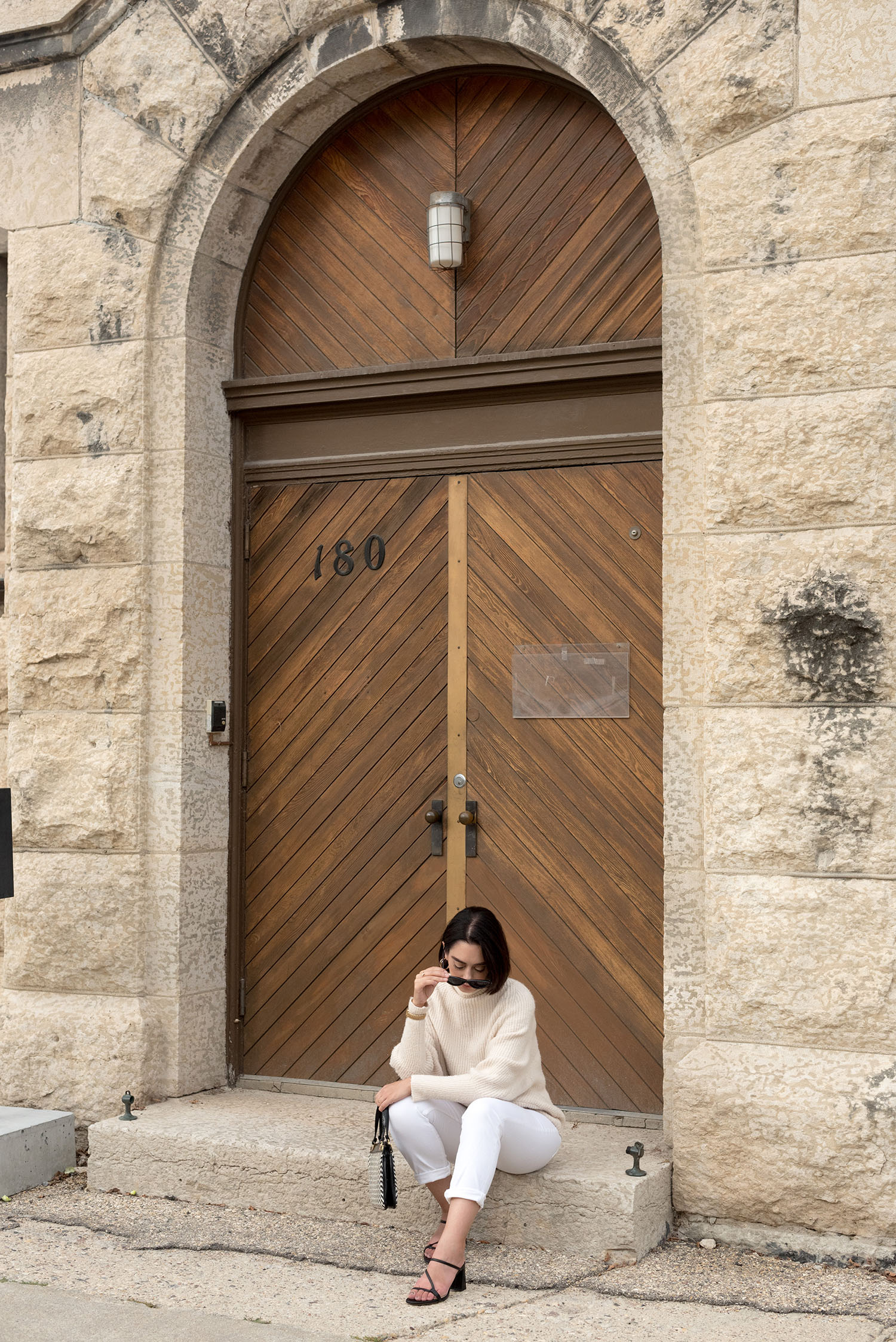 Top Winnipeg fashion blogger Cee Fardoe of Coco & Vera sits outside a church wearing white Mango jeans and Zara strappy sandals