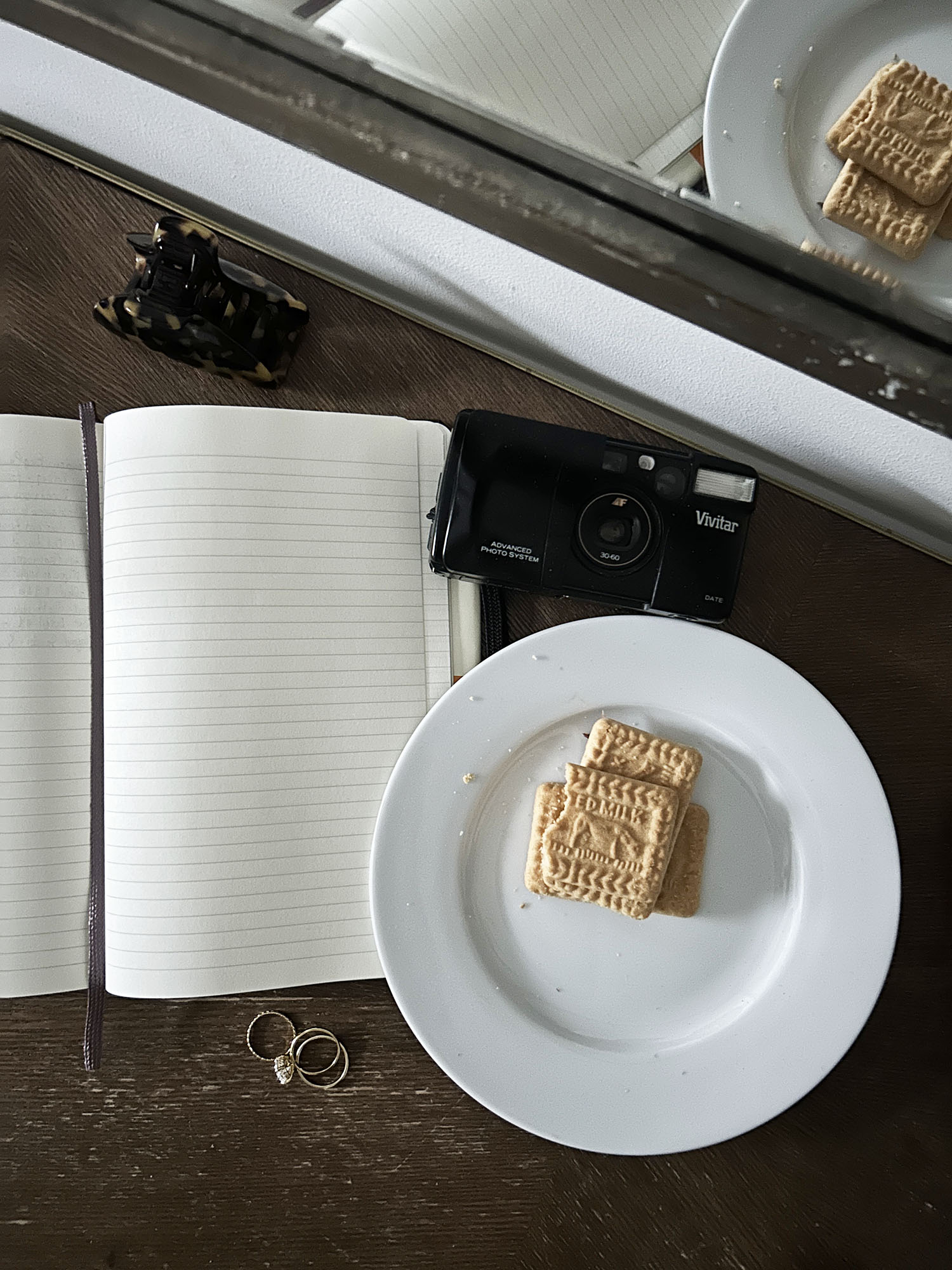 Coco & Voltaire - Moleskine notebook, plate of butter biscuits, vintage camera, rings and hair clip on a wooden table